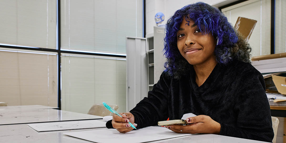 Image of an MGA student smiling while sitting at a table with a phone in one hand and a writing utensil in the other.